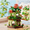 A miniature building set featuring a green woodland cabin with orange roses and pink flowers, displayed on a wooden surface.