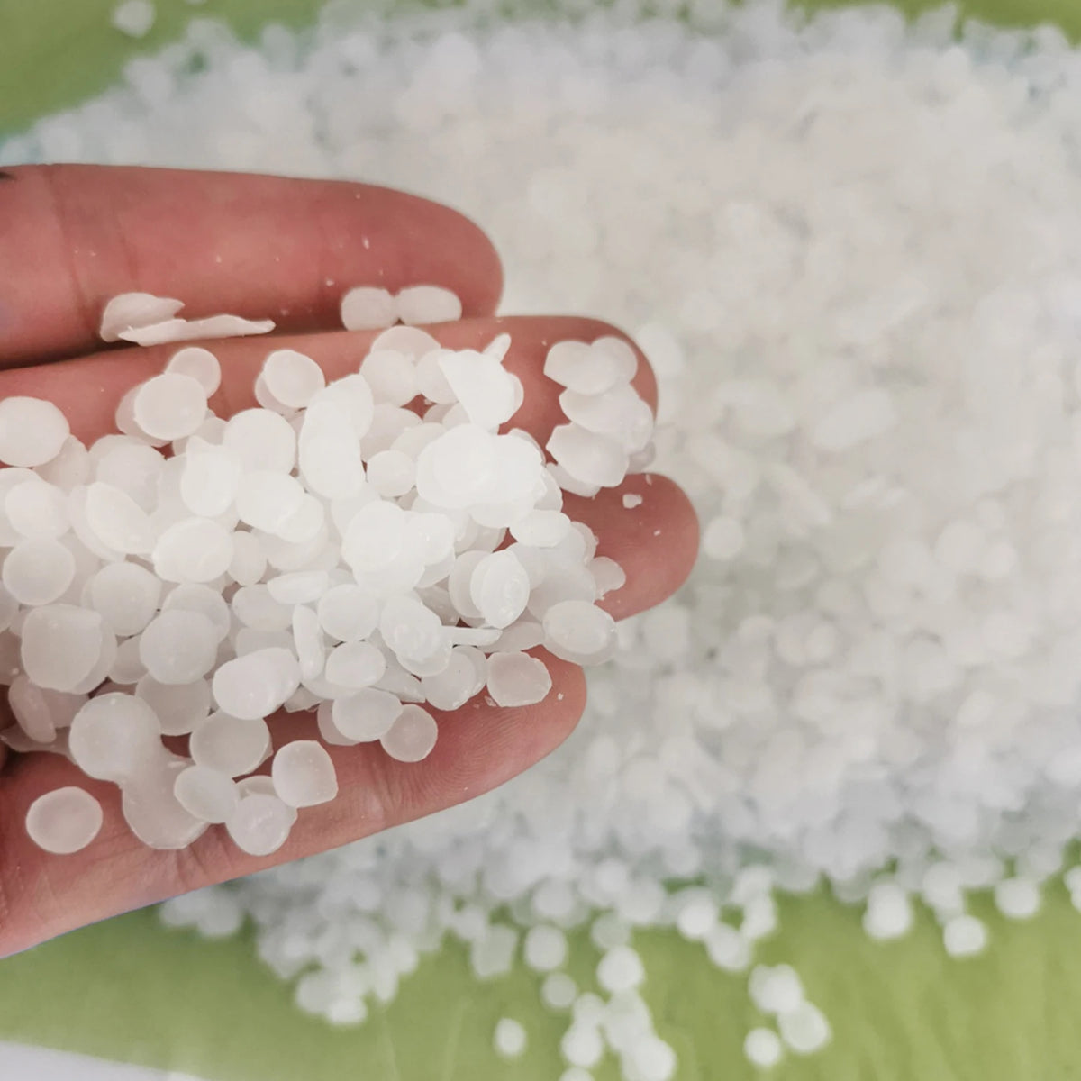A hand holds small white wax pellets next to a pile of natural beeswax for DIY candle making and skincare.