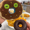 A hand holds a plush toy resembling an ugly cute monster donut with large green eyes and brown body, next to a dark chocolate