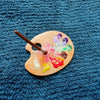 A wooden palette with paint on it and a paintbrush resting on the edge, set against a dark blue textured fabric.
