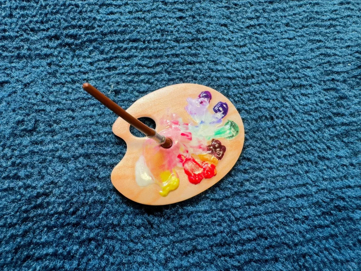 A wooden palette with paint on it and a paintbrush resting on the edge, set against a dark blue textured fabric.