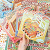 A hand holds a colorful sticker book featuring various shop displays and scenes for DIY crafts.