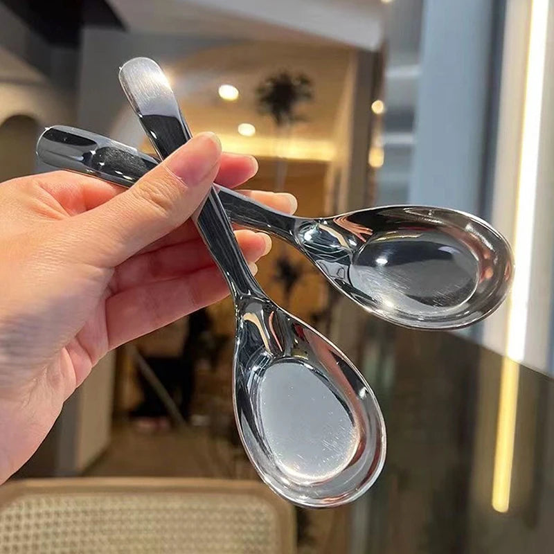 A hand holds a clear glass spoon and fork set against a blurred indoor background.