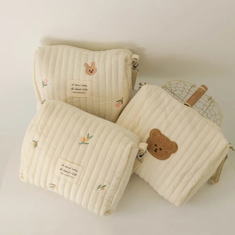 A cream-colored quilted cotton diaper and organizer bag with a small bear charm.