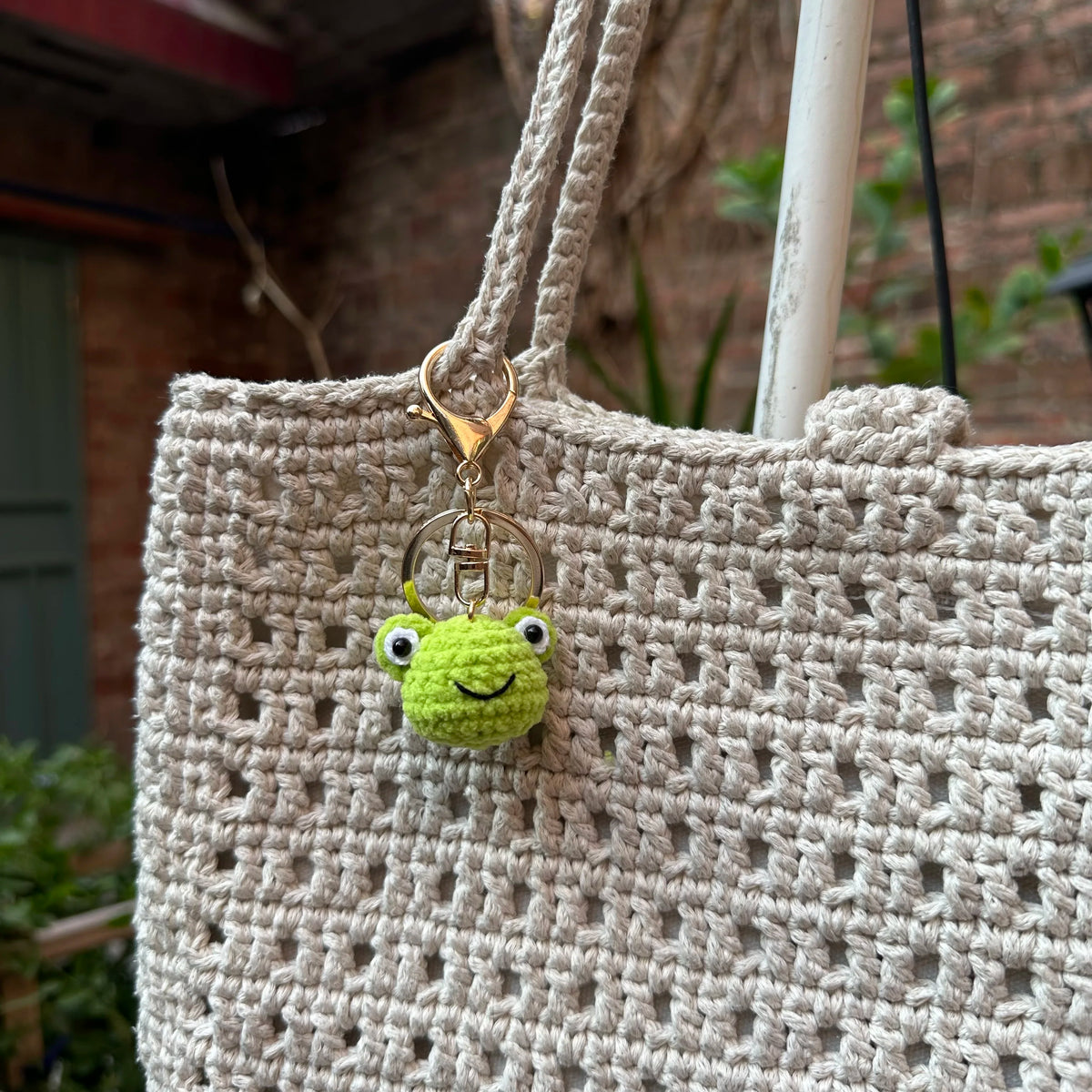 A light beige crocheted bag with a gold-colored keychain featuring a small green rabbit or chick charm.