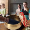 A handcrafted Tibetan singing bowl with a gold-colored bowl, silk cushion, and wooden mallet resting on a dark surface.