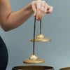A hand holds a brass tingsha cymbal suspended by a dark cord above another brass cymbal.