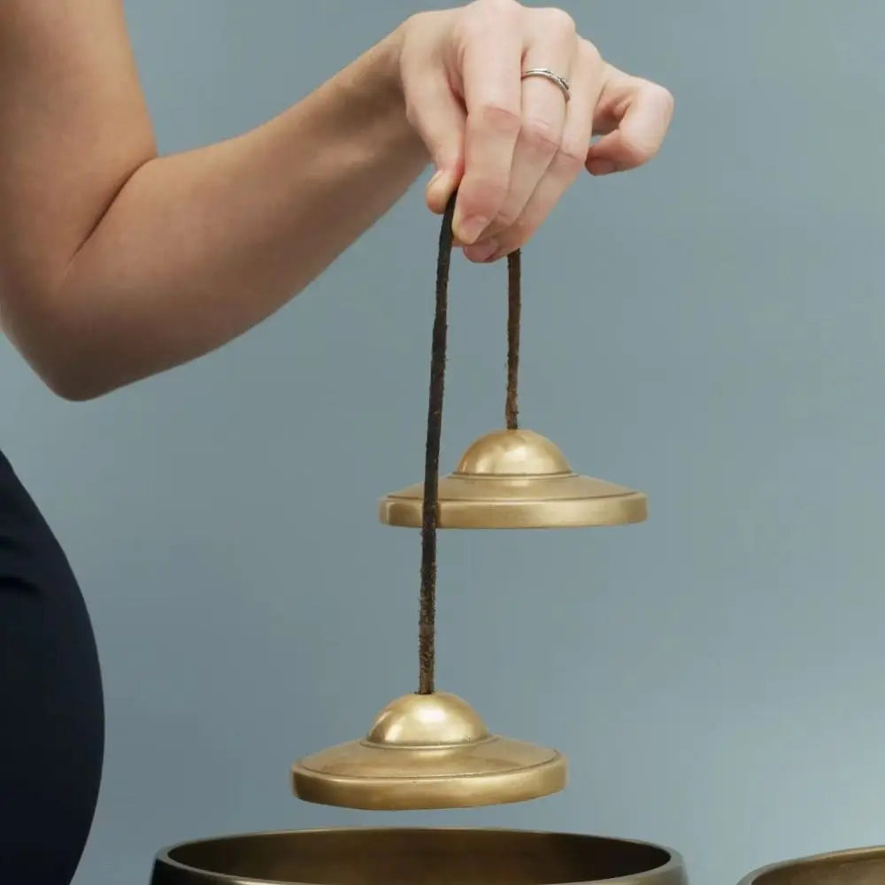 A hand holds a brass tingsha cymbal suspended by a dark cord above another brass cymbal.