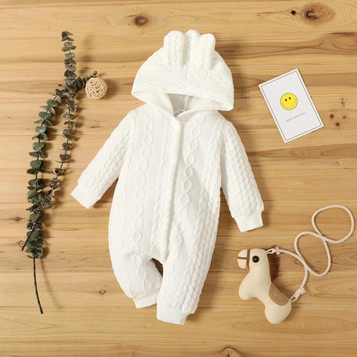 A white knitted hooded baby romper lying on a wooden surface with a small yellow smiley face card and a wooden toy.