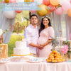 A pregnant woman and man stand together in a pink and gold setting for a gender reveal party decoration display.
