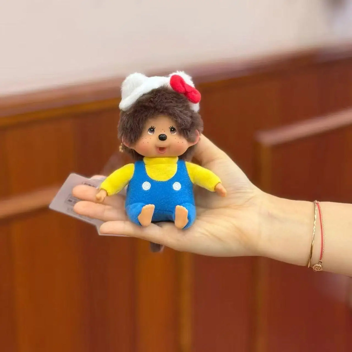 A hand is holding a Monchhichi plush monkey with a white and red headband against a wooden background.