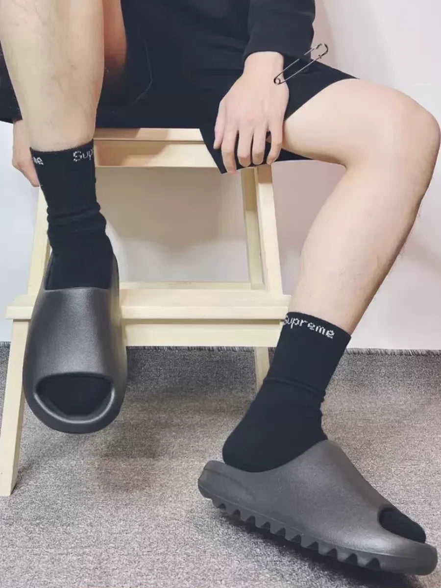 A person wearing black socks and grey EVA platform slides is sitting on a wooden stool.