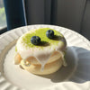 A round soufflé with white glaze and green sprinkles is topped with two blueberries on a white plate.