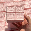 A hand holds a pink strawberry milk squishy stress relief toy against a grid of similar squishies.