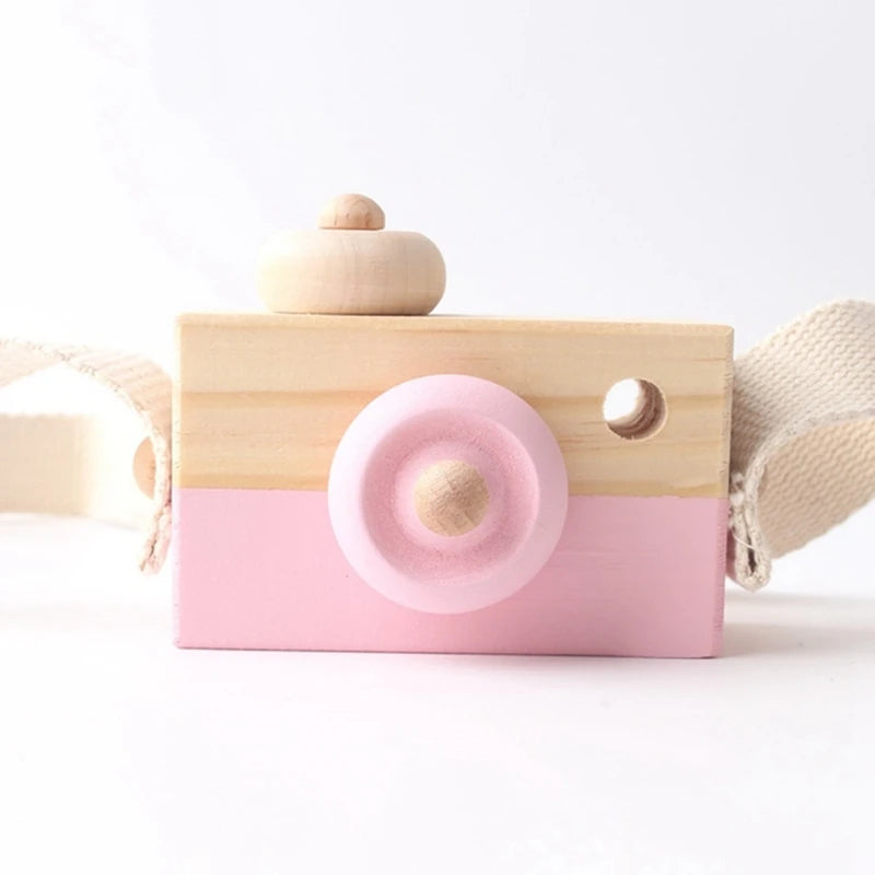 A pink wooden camera teether toy with a round lens and a wooden handle is shown against a white background.
