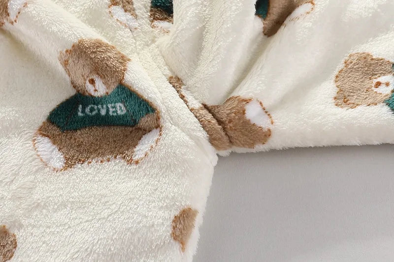 A close-up of a cream fleece jacket and pants set featuring brown bear cartoon prints with green accents.