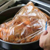 A hand is opening a clear heat-resistant roasting bag filled with seasoned meat, likely turkey or chicken, inside an oven pan