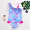 A purple and blue mermaid unicorn one-piece swimsuit with ruffled trim, displayed next to a green leaf and sunglasses.