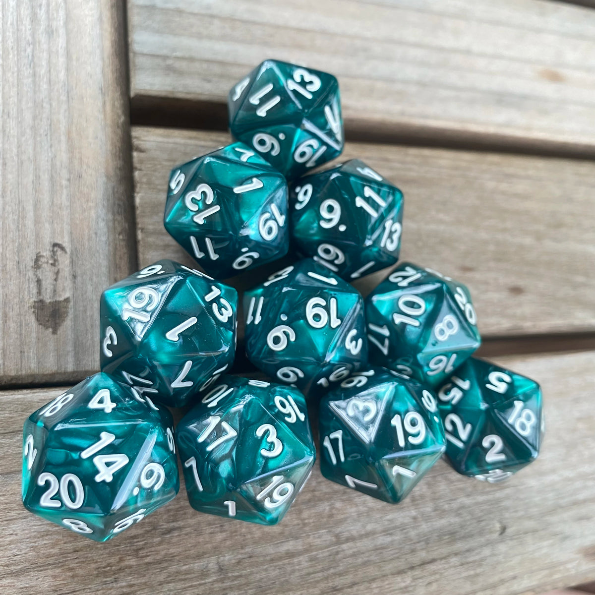 A pile of ten teal acrylic D20 dice with various numbered faces resting on a wooden surface.