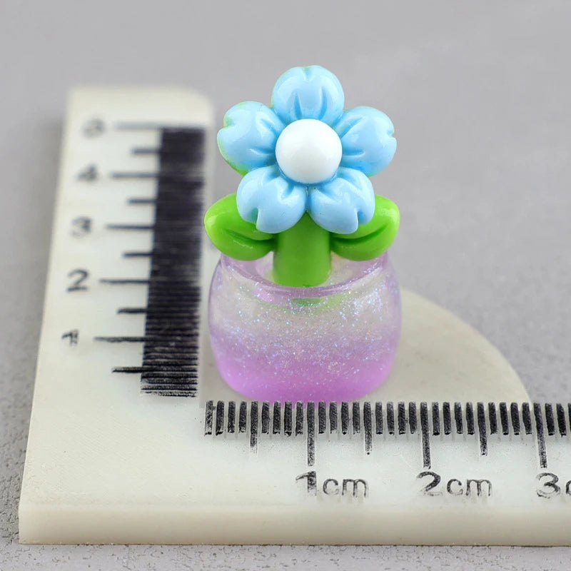 A miniature blue flower with a white center is placed in a small green pot on a ruler.