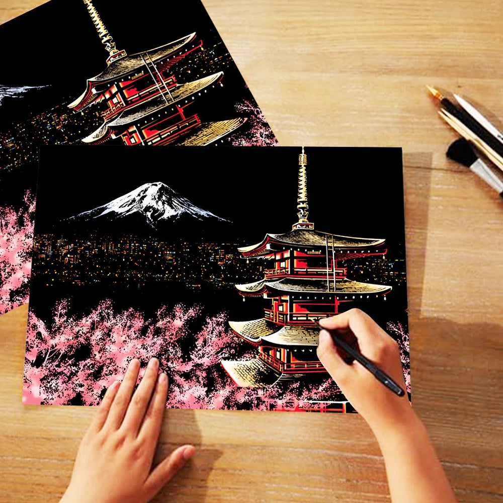 A hand is painting a scratch art scene featuring a Japanese pagoda and Mount Fuji against a night sky background.