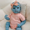 A lifelike reborn baby doll with blue skin and pink clothing is resting on a white fluffy surface.