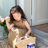 A young girl wearing a beige sleeveless cotton sundress sits outdoors holding a woven basket labeled Porto
