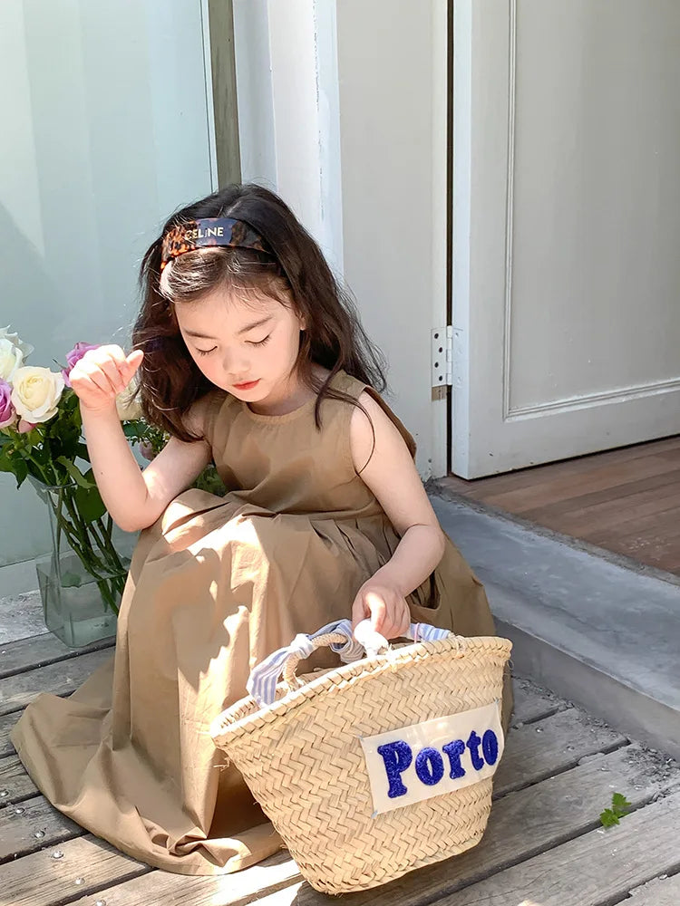 A young girl wearing a beige sleeveless cotton sundress sits outdoors holding a woven basket labeled Porto