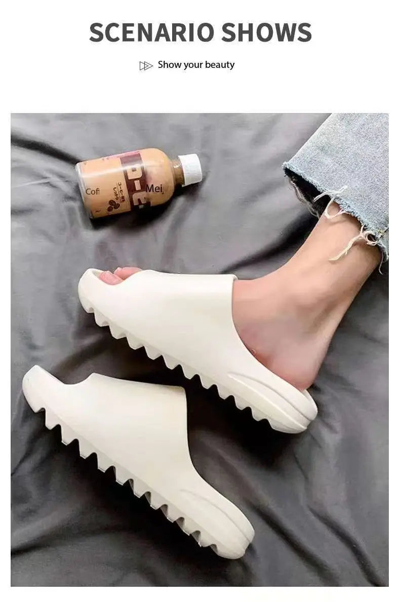 A pair of white EVA platform slides shown next to a bottle of oil on a dark gray fabric.