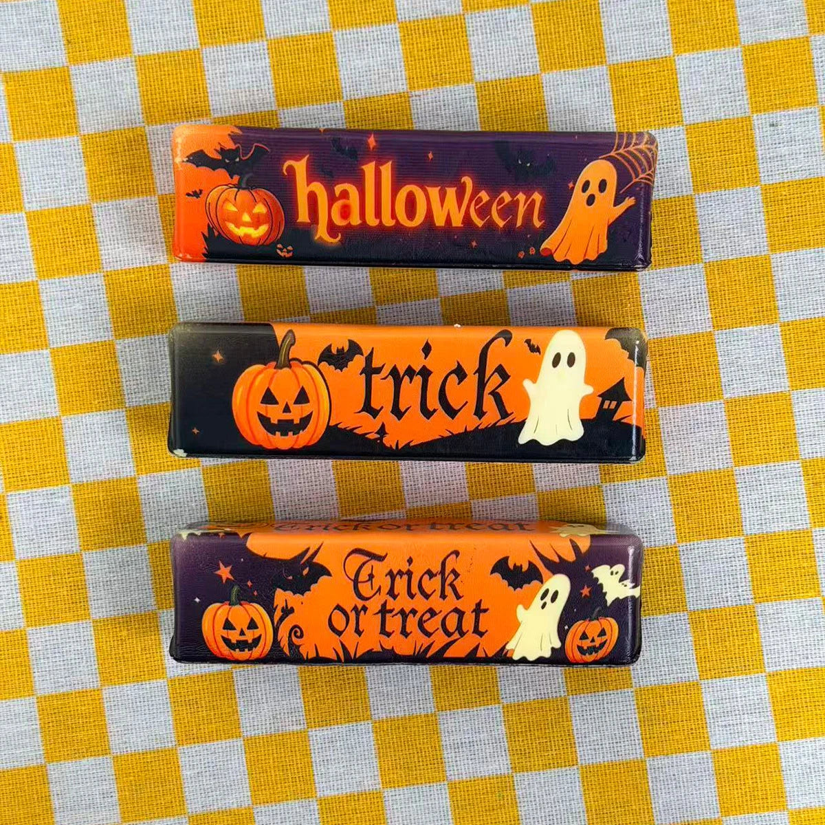 Three Halloween themed butter squishy toys featuring pumpkins and ghosts on a yellow checkered background.