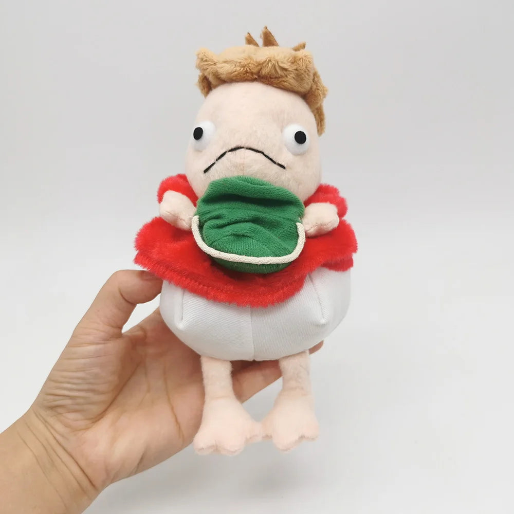 A light pink plush toy of a hippo with brown hair, wearing a red cape and green collar, being held by a hand.