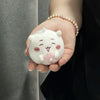 A small white plush charm shaped like a bear face with pink details is held in a person's hand.