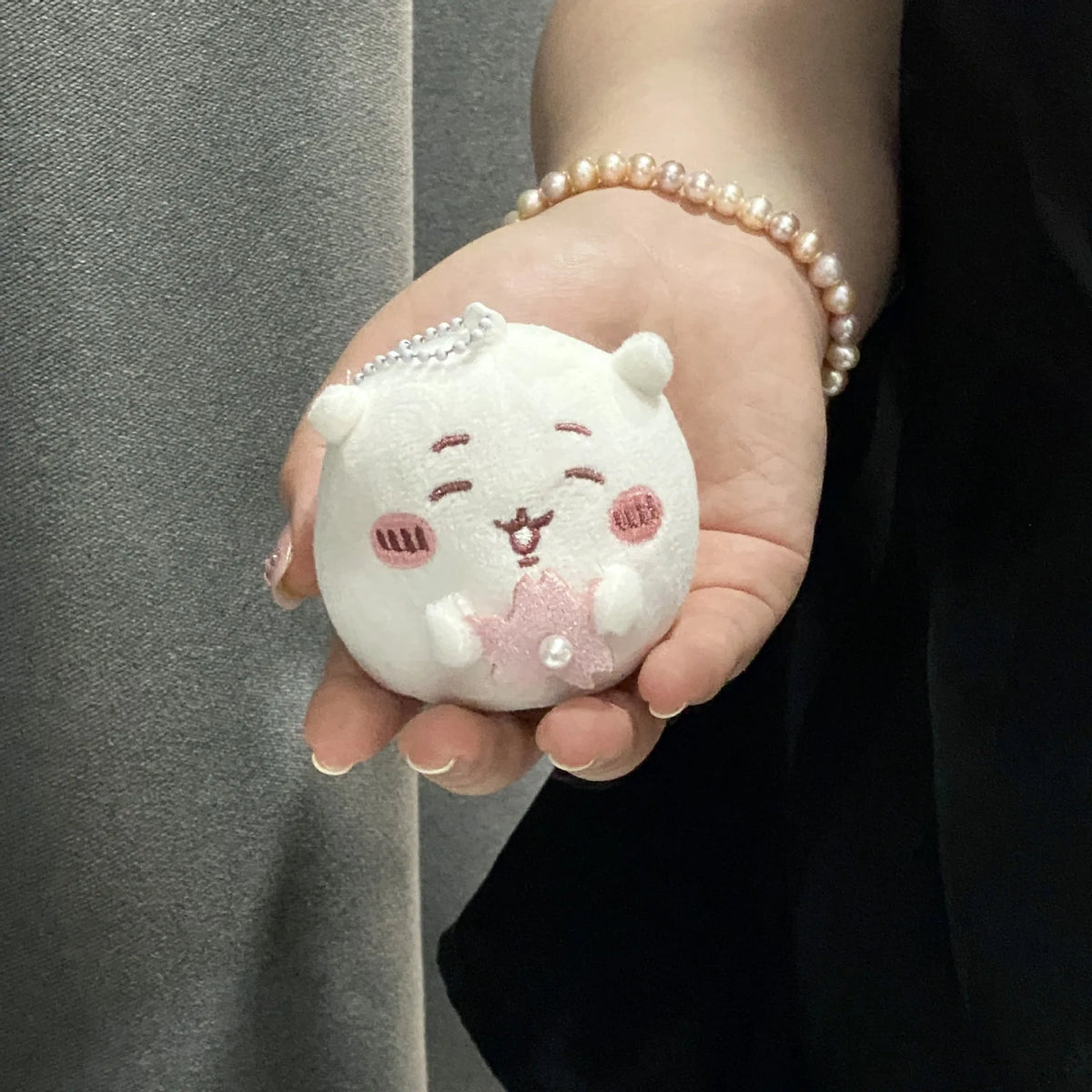 A small white plush charm shaped like a bear face with pink details is held in a person's hand.
