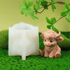 A small, pink silicone candle mold of a Highland cow sits on green moss with a branch of leaves above it.
