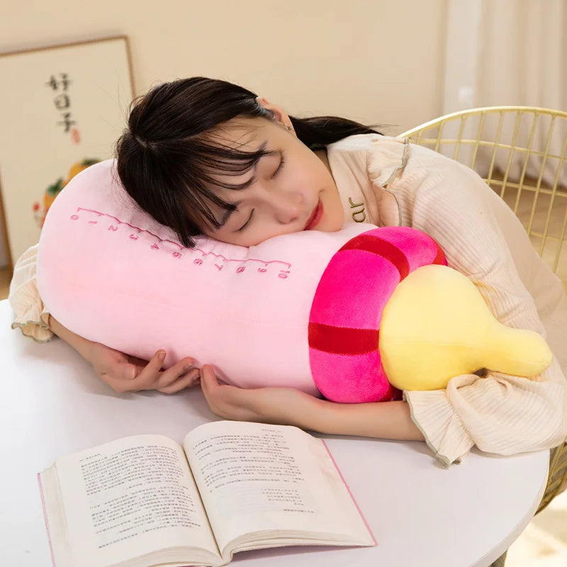 A person is resting their head on a pink plush pillow shaped like a milk bottle, with a yellow cushion attached, indoors.