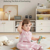 A baby wearing a pink sleeveless sleeping bag is sitting on a rug in a bright room.