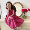 A young girl wearing a pink sleeveless cotton sundress is sitting in front of a wicker mirror.