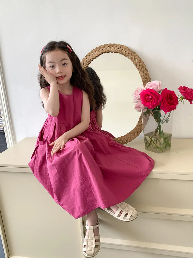 A young girl wearing a pink sleeveless cotton sundress is sitting in front of a wicker mirror.