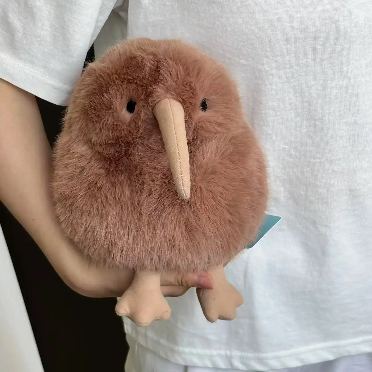 A fluffy, brown kiwi bird plush toy is held by a person against a white shirt.