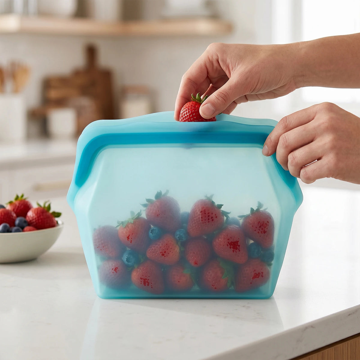 A hand is placing a strawberry into a blue silicone storage bag filled with fresh berries on a white countertop.