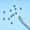 A silver precision bead and jewelry pick-up tool with a textured handle and several small silver beads scattered on a light b