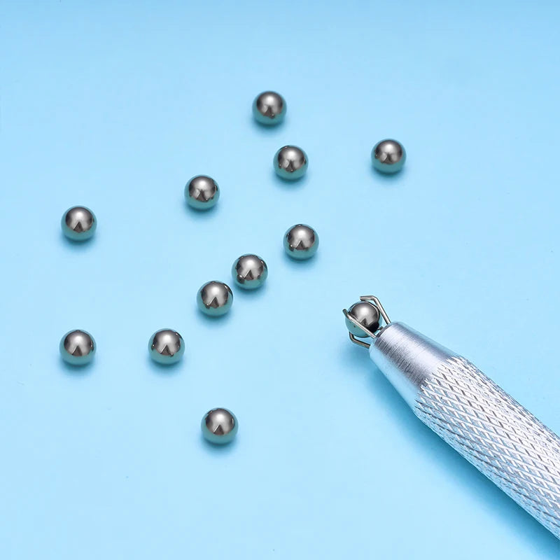 A silver precision bead and jewelry pick-up tool with a textured handle and several small silver beads scattered on a light b