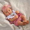 A lifelike vinyl reborn doll with a pink floral headband and a red and white checkered outfit lying on a cream blanket.