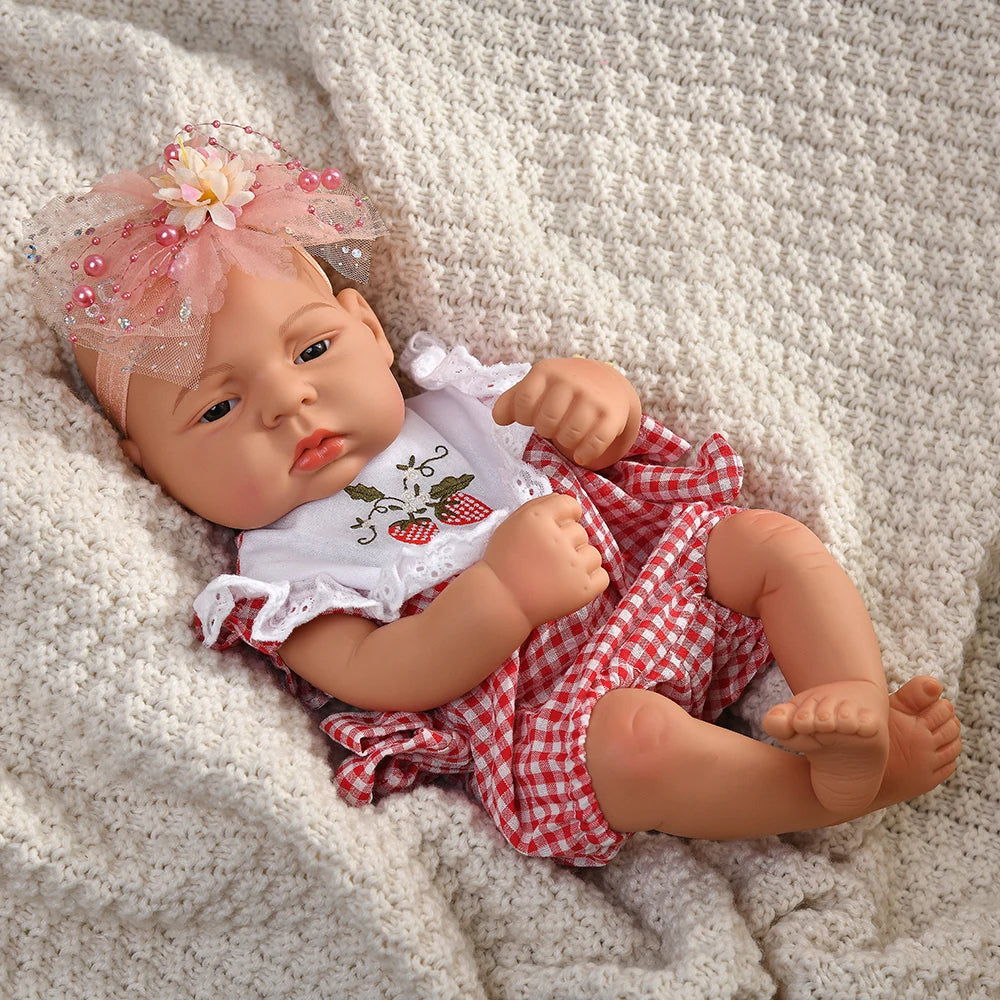 A lifelike vinyl reborn doll with a pink floral headband and a red and white checkered outfit lying on a cream blanket.