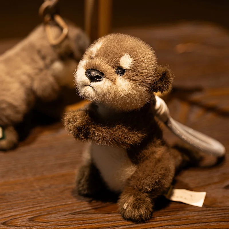 A small, brown plush otter charm resting on a wooden surface next to a larger stuffed animal.