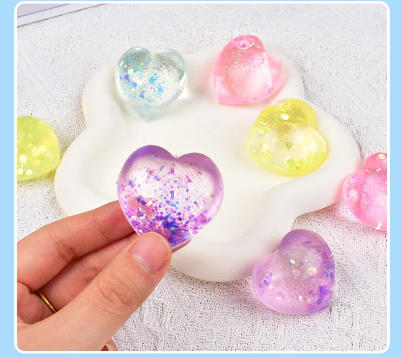 A hand holds a white heart-shaped squishy toy filled with glittery, translucent foam in various colors.
