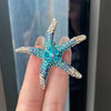 A hand holds a sparkling blue and gold rhinestone turtle brooch pin against a dark background.
