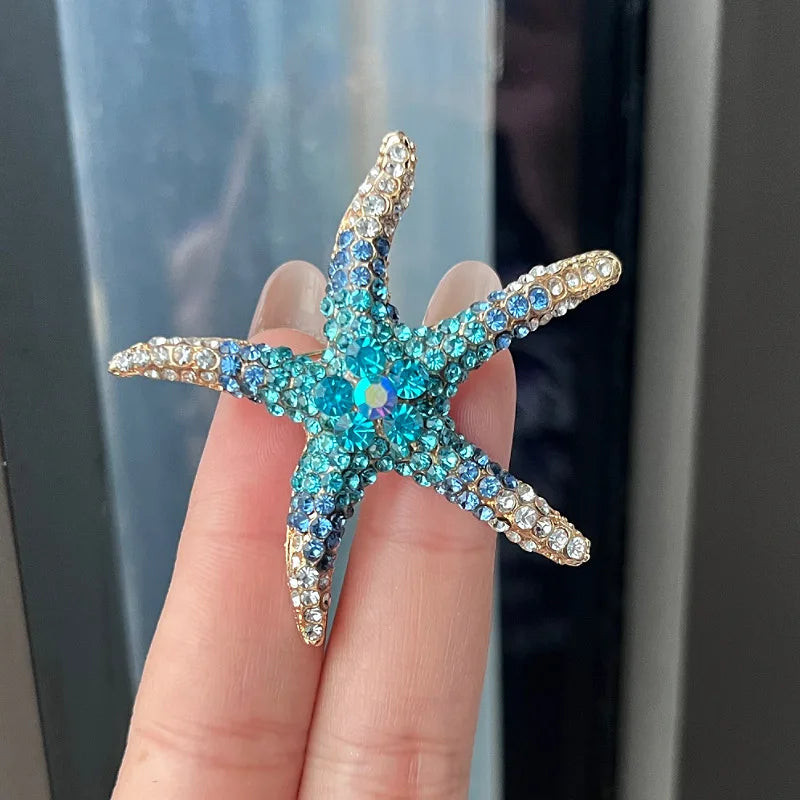 A hand holds a sparkling blue and gold rhinestone turtle brooch pin against a dark background.
