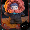 A fluffy, reddish-brown plush doll with purple hair and blue accents sits on a wooden surface.