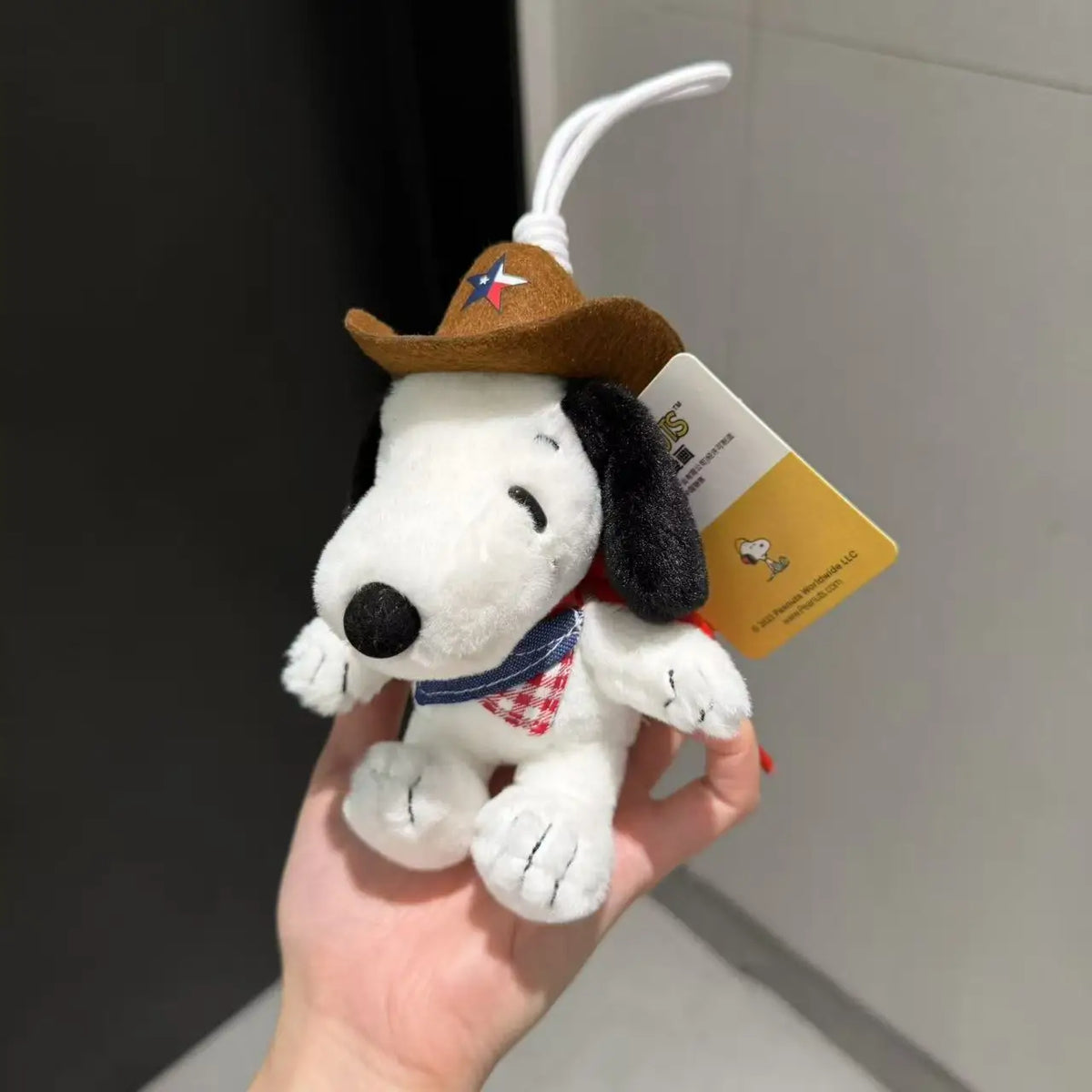 A white dog plush toy wearing a brown cowboy hat and a red and white plaid shirt, attached to a white string for a keychain.